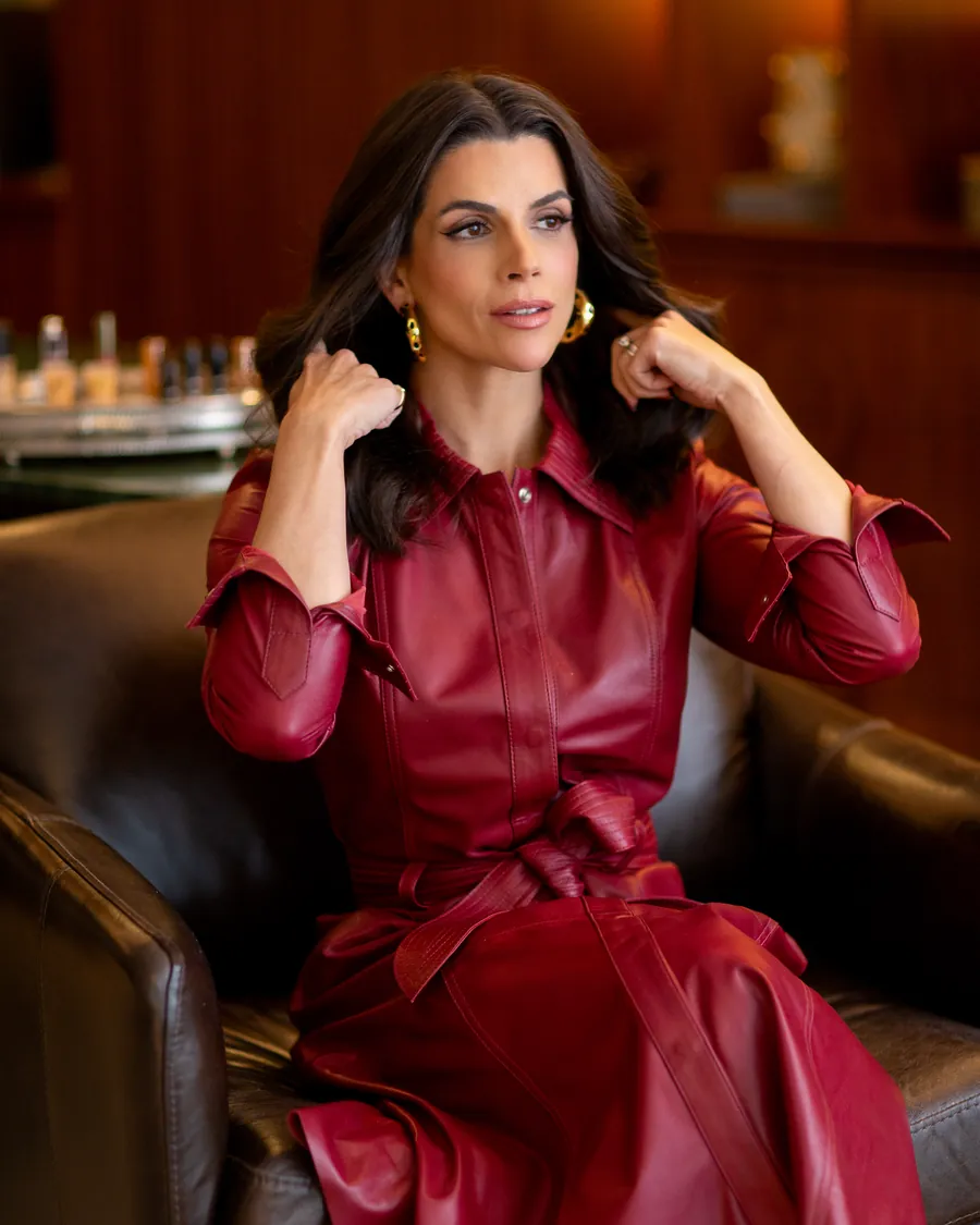 Mel Galhardo sentada em poltrona de couro, de vestido bordô, com a mão no cabelo, sorrindo