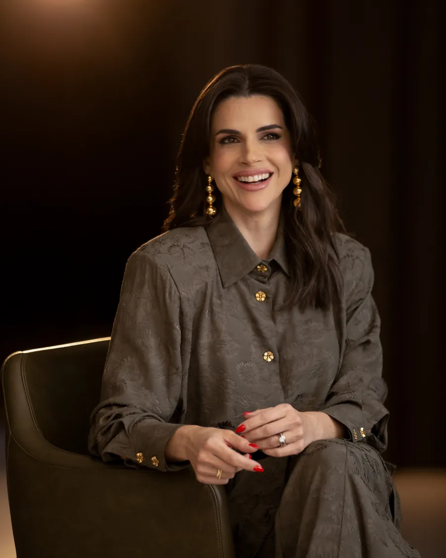 Mel Galhardo sentada, de blusa cinza, sorrindo, em ambiente com luz quente