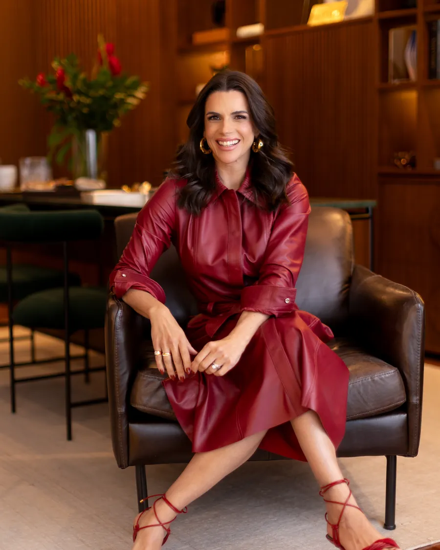 Mel Galhardo de vestido de couro bordô, sentada em poltrona no seu closet, pronta para uma consultoria de imagem