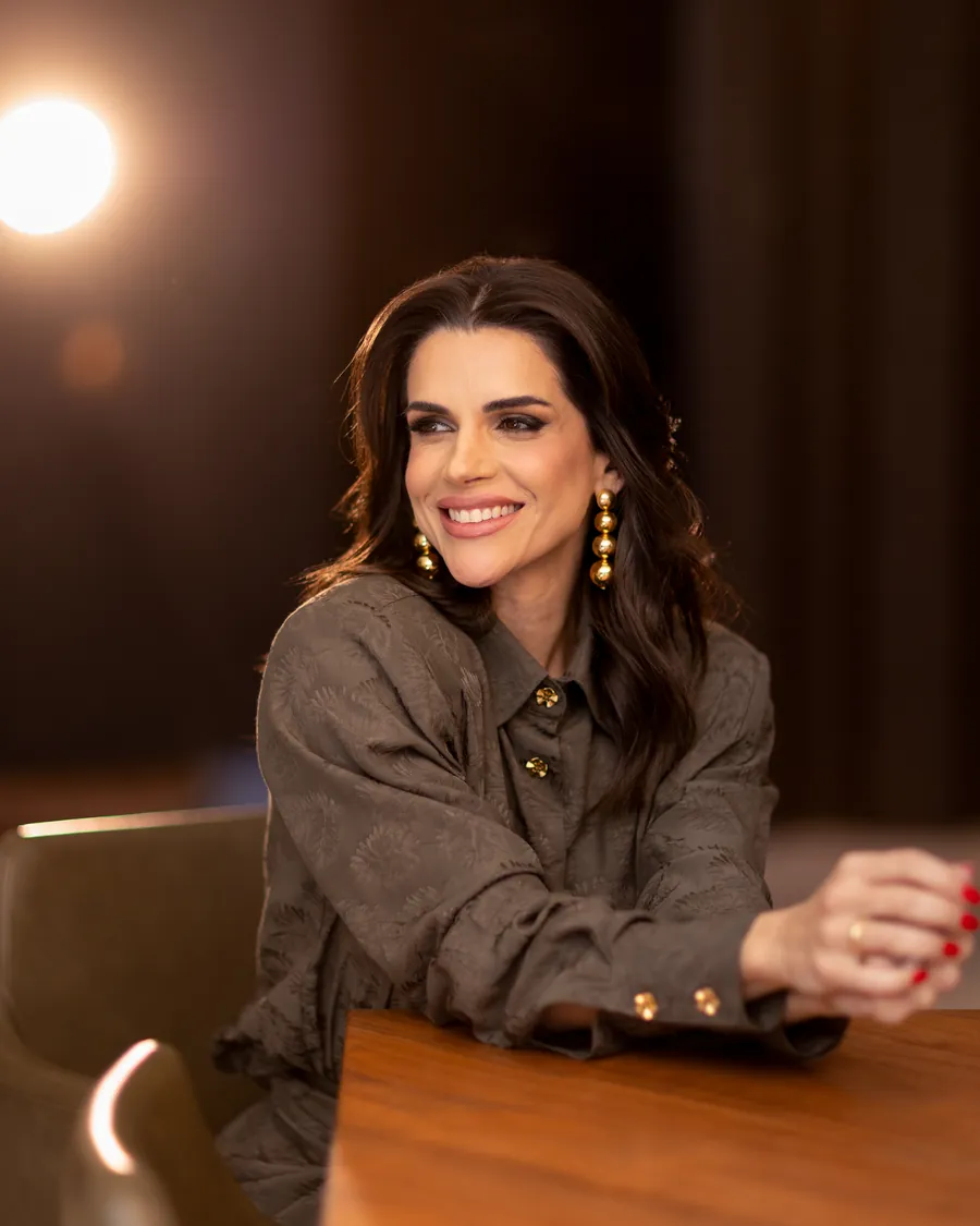 Mel Galhardo sentada à mesa, de blusa cinza, sorrindo, com as mãos apoiadas