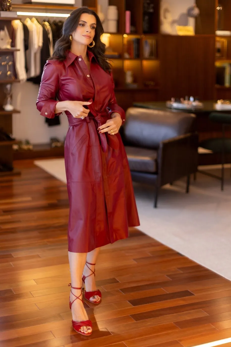 Mel Galhardo de corpo inteiro no ateliê, de vestido vinho, com araras de roupas ao fundo