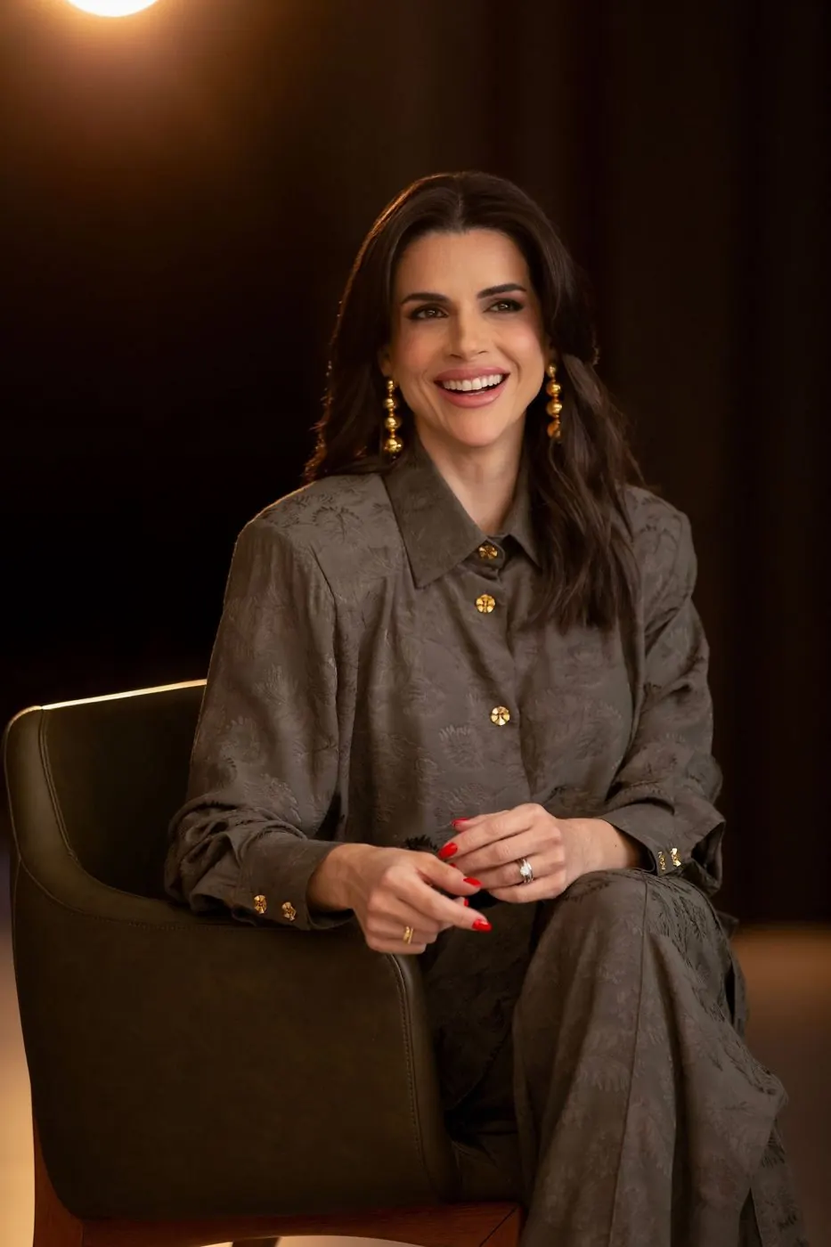 Mel Galhardo sorrindo, sentada em uma poltrona, com brincos dourados