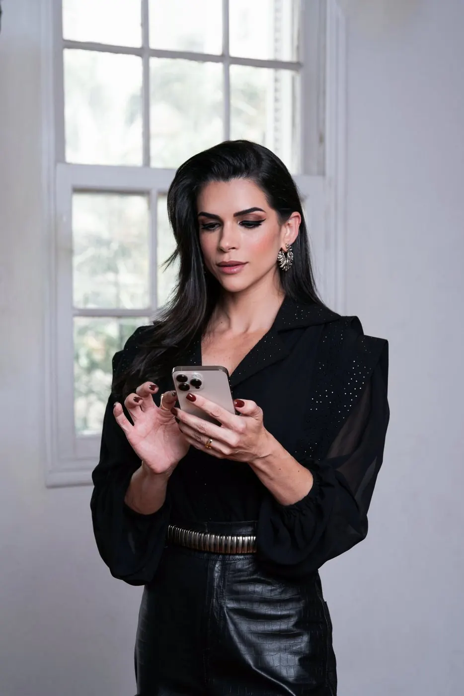 Mel Galhardo de preto, olhando o celular junto à janela